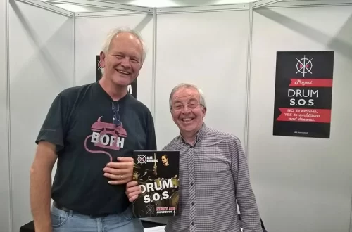 Aleš Korošec z gosti na London Drum Show, ob promociji priročnika Bobnarski S.O.S. (DRUM S.O.S). V rokah je izvod priročnika, v ozadju pa plakat istoimenskega projekta.