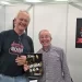 Aleš Korošec z gosti na London Drum Show, ob promociji priročnika Bobnarski S.O.S. (DRUM S.O.S). V rokah je izvod priročnika, v ozadju pa plakat istoimenskega projekta.