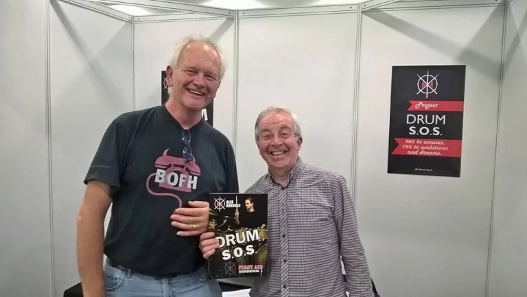 Aleš Korošec z gosti na London Drum Show, ob promociji priročnika Bobnarski S.O.S. (DRUM S.O.S). V rokah je izvod priročnika, v ozadju pa plakat istoimenskega projekta.